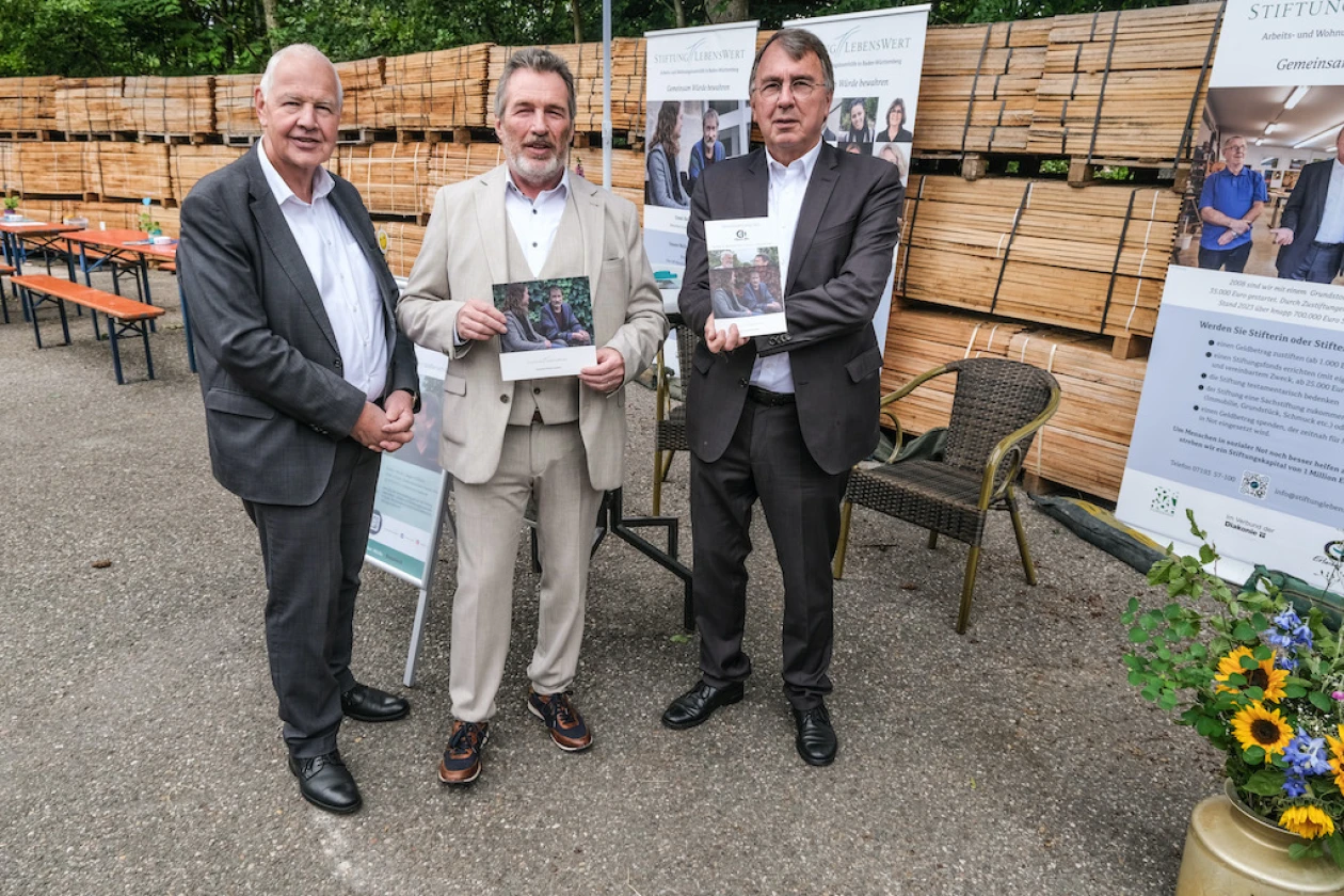 Drei M&auml;nner stehen vor einem Stehtisch und pr&auml;sentieren Brosch&uuml;ren.