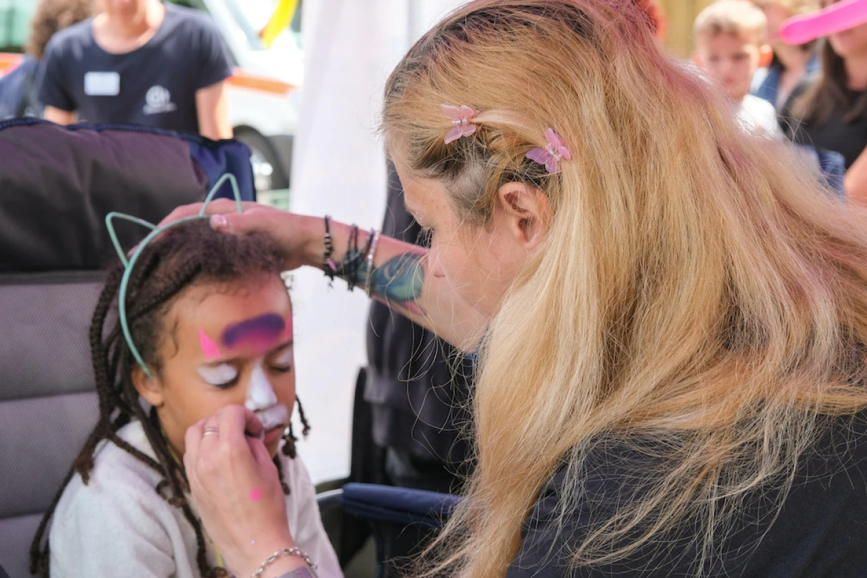 Eine Visagistin bemalt das Gesicht eines Kindes mit bunten Schminken in Pastellfarben bei einem Fest. Die Frau tr&auml;gt blondes Haar und konzentriert sich sorgf&auml;ltig auf die Gesichtsmalerei, w&auml;hrend das Kind still sitzt.