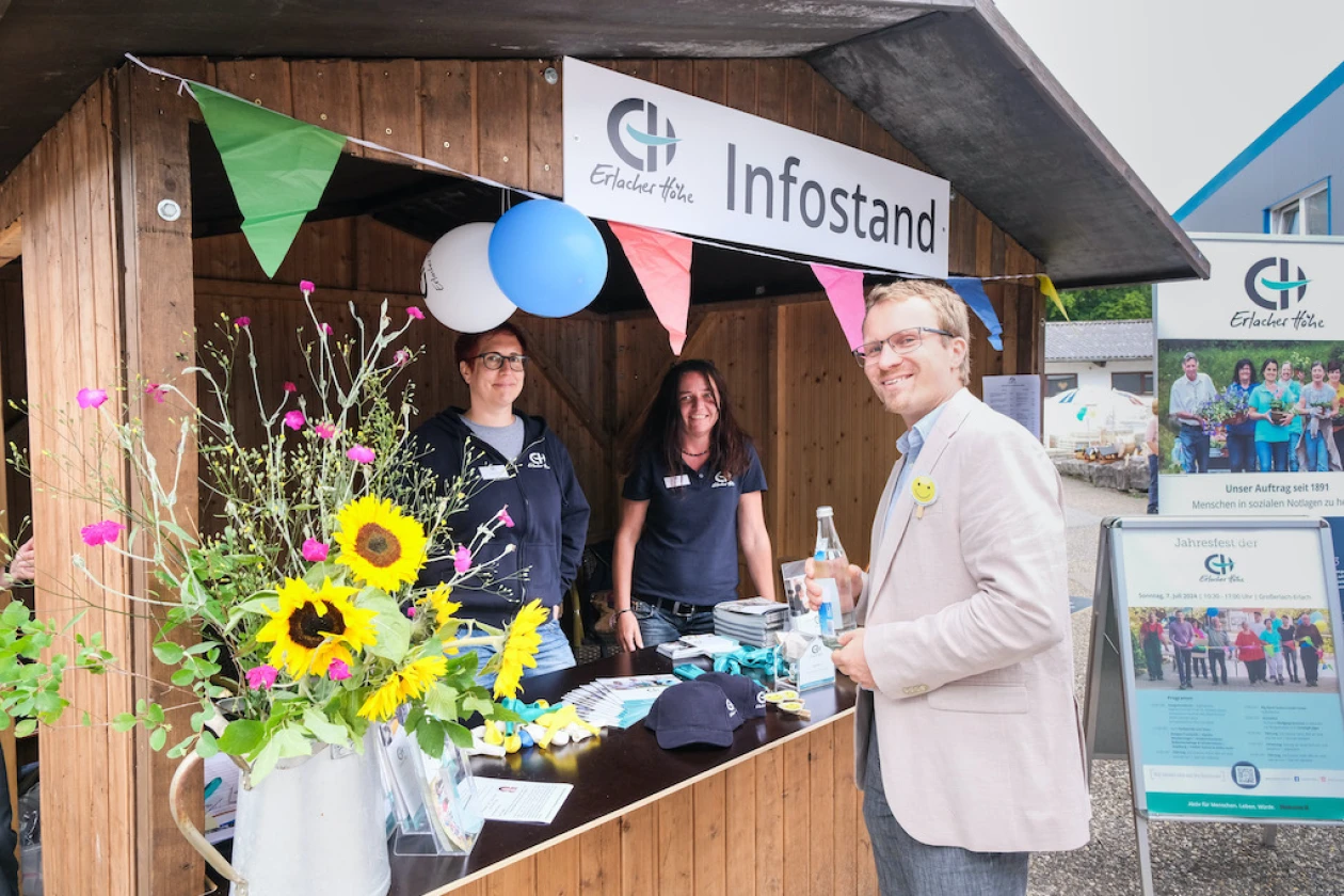 Zwei Frauen in dunklem Oberteil mit Logo der Erlacher H&ouml;he stehen an einem Infostand mit Sonnenblumenstrau&szlig;, bunten Wimpeln, blauen Luftballons und Plakataufsteller. Ein Mann steht davor und alle l&auml;cheln in die Kamera. Die Holzh&uuml;tte tr&auml;gt die Aufschrift "Infostand".