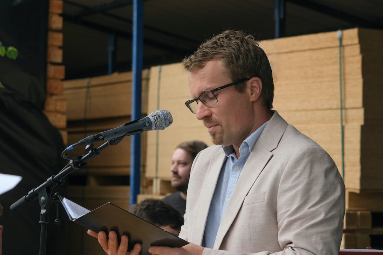 Ein Mann in hellblauem Hemd und beigem Sakko steht auf einer B&uuml;hne vor einem Mikrofon und h&auml;lt eine Mappe in der Hand. Es scheint als liest er vor.