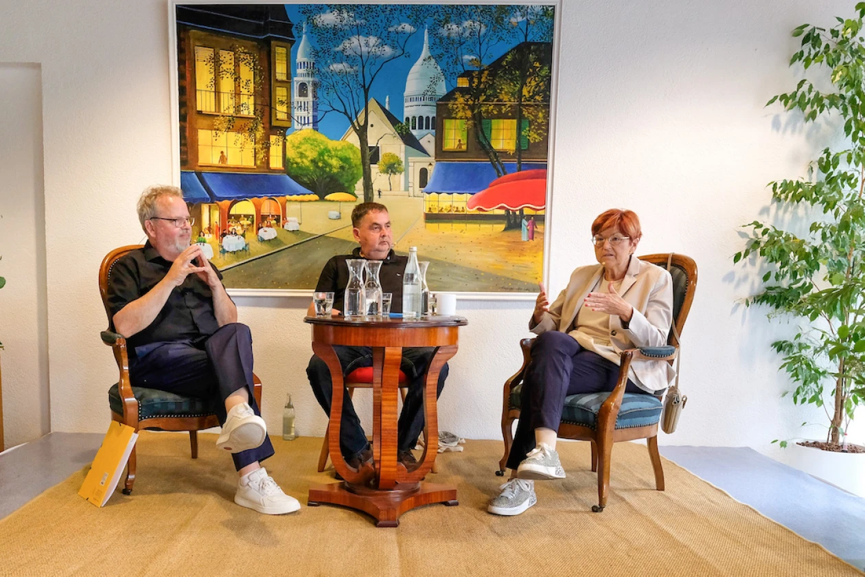 Drei Personen sitzen auf St&uuml;hlen um einen kleinen Tisch in einer Gespr&auml;chssituation