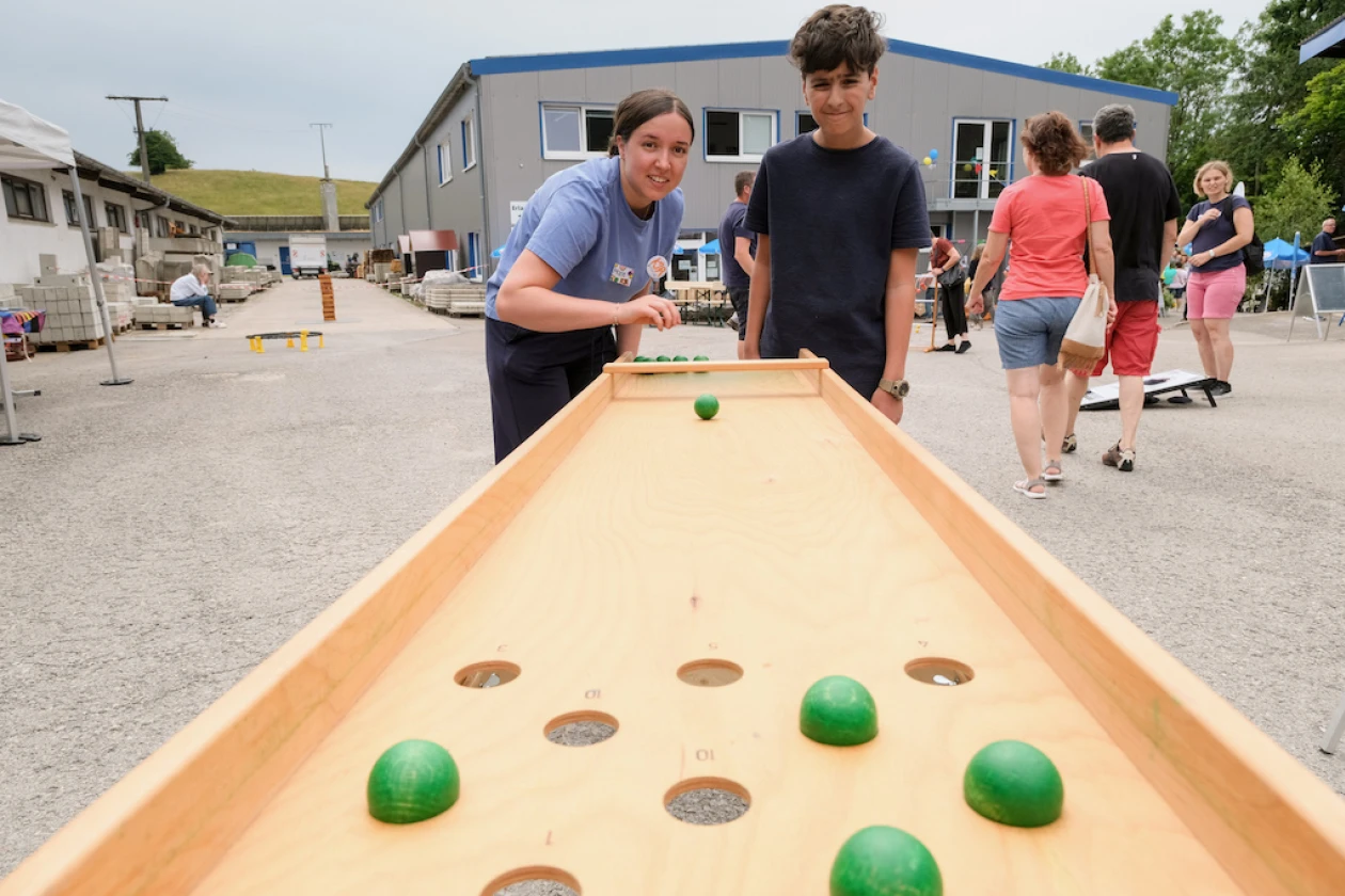 Ein Junge und eine Frau spielen zusammen auf einer Holzbahn mit gr&uuml;nen Kugeln w&auml;hrend eines Sommerfests auf dem Gel&auml;nde der Erlacher H&ouml;he, im Hintergrund sind weitere Besucher zu sehen.