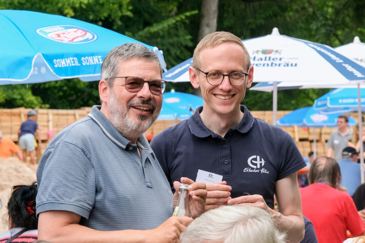Zwei M&auml;nner l&auml;cheln in die Kamera, w&auml;hrend sie zusammen ein St&uuml;ck Kuchen halten