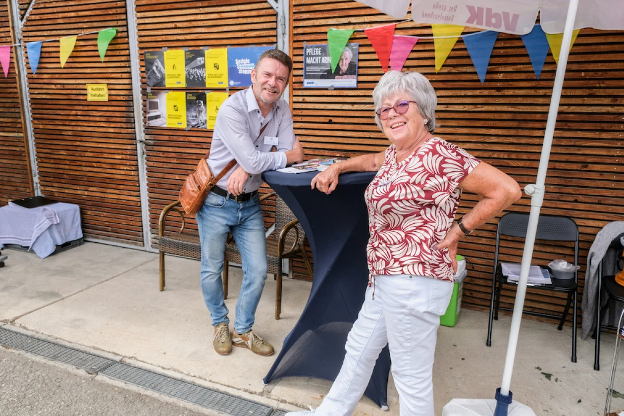 Zwei Personen stehen an einem Stehtisch bei einem Fest. Im Hintergrund sind bunte Girlanden und Dekoration an einer Holzwand zu sehen.