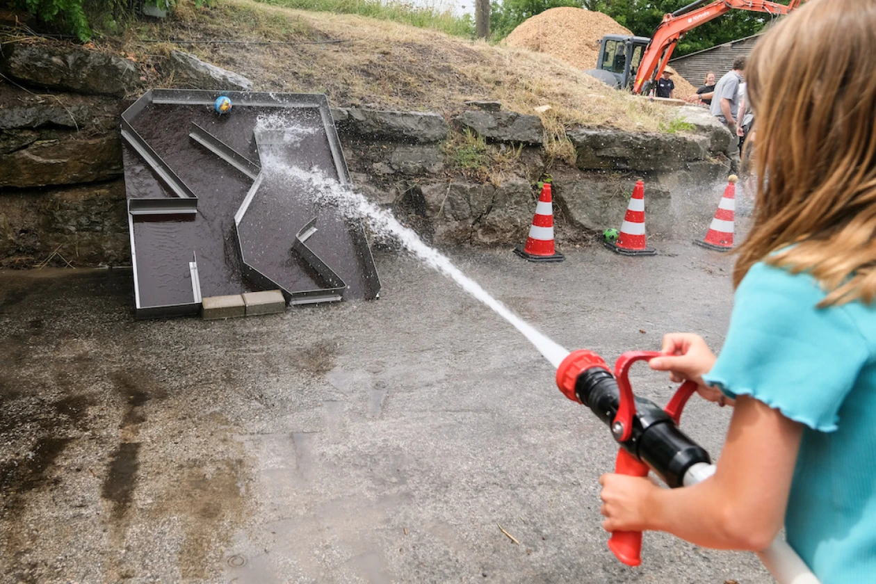 Ein Kind in blauem Shirt h&auml;lt einen roten Feuerwehrschlauch und spr&uuml;ht Wasser