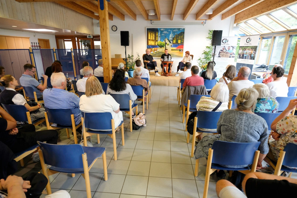 Ein Saal mit heller Holzdecke und gro&szlig;en Fenstern ist mit Besuchern gef&uuml;llt, die auf blauen St&uuml;hlen sitzen und nach vorne zu einer B&uuml;hne blicken.