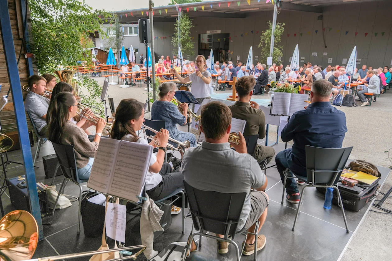 Musikensembles und Besucher sitzen in einem Schulhof w&auml;hrend eines Jahresfests. Im Vordergrund spielen Musiker mit Noten, im Hintergrund sind zahlreiche Zuschauer.