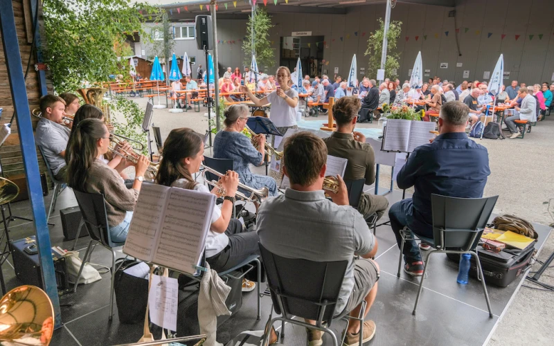 Musikensembles und Besucher sitzen in einem Schulhof w&auml;hrend eines Jahresfests. Im Vordergrund spielen Musiker mit Noten, im Hintergrund sind zahlreiche Zuschauer.