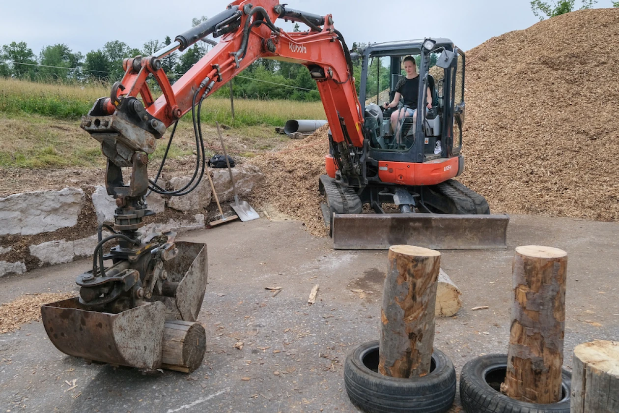 Ein roter Baggerarm mit Greifer bearbeitet Holzst&auml;mme auf einem Lagerplatz, w&auml;hrend ein Bediener in der Kabine des Baggers sitzt