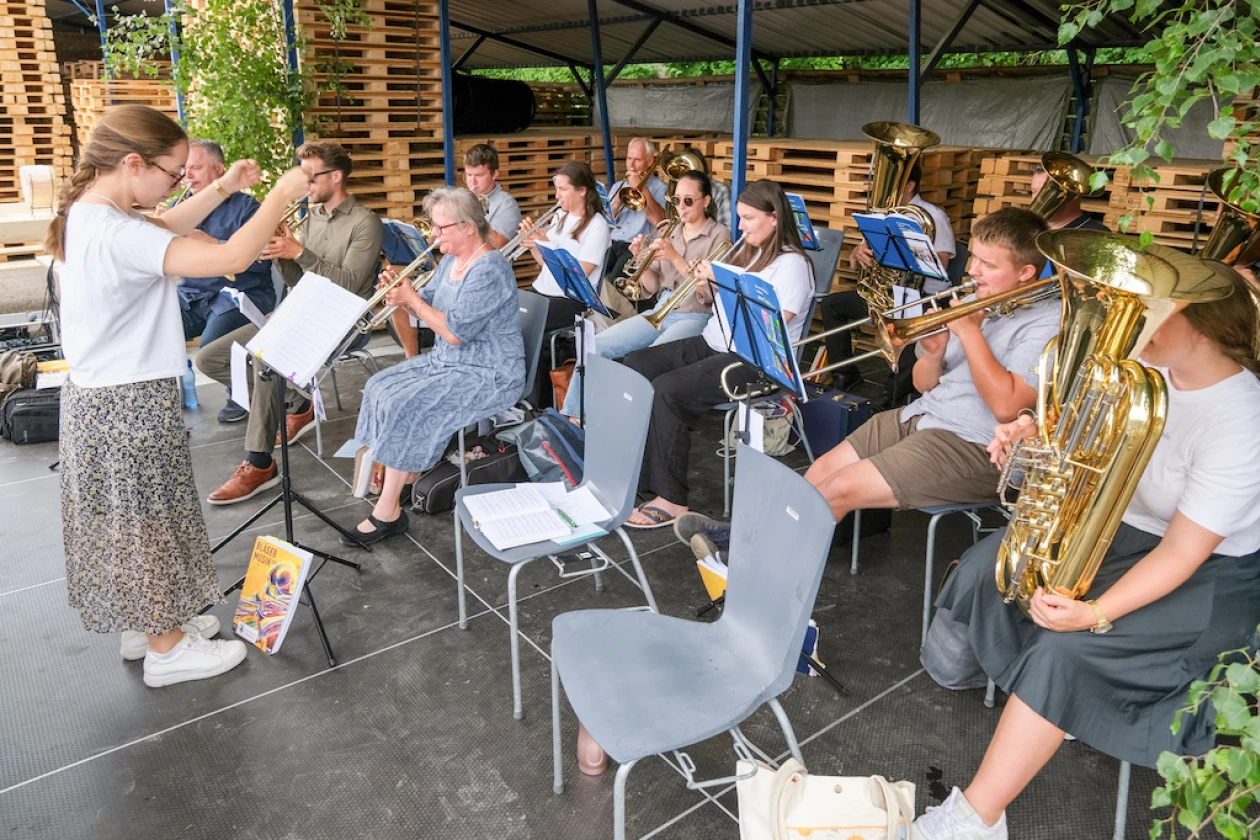 Eine Gruppe von Musikern spielt zusammen in einem Au&szlig;enbereich. Die Musiker sitzen auf St&uuml;hlen und spielen verschiedene Instrumente, haupts&auml;chlich Blechbl&auml;ser wie Trompeten und Posaunen