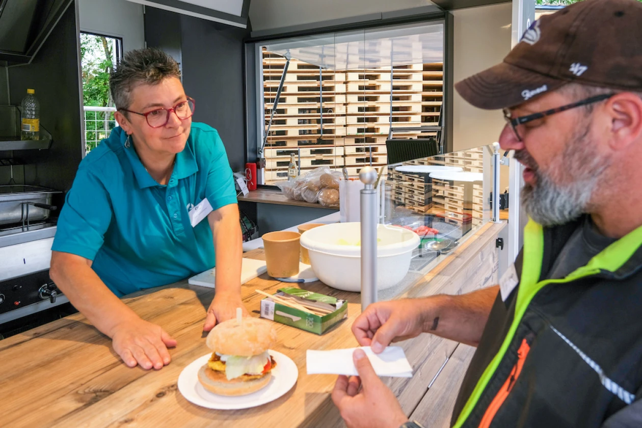 Eine Frau in blauem Poloshirt und Brille steht hinter einer K&uuml;chentheke und reicht einem Mann mit Kappe einen Teller mit einem Burger