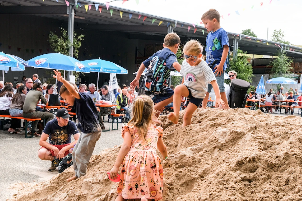 Kinder spielen auf einem Sandh&uuml;gel w&auml;hrend Erwachsene unter blauen Sonnenschirmen im Hintergrund an Tischen sitzen und das Jahresfest feiern.