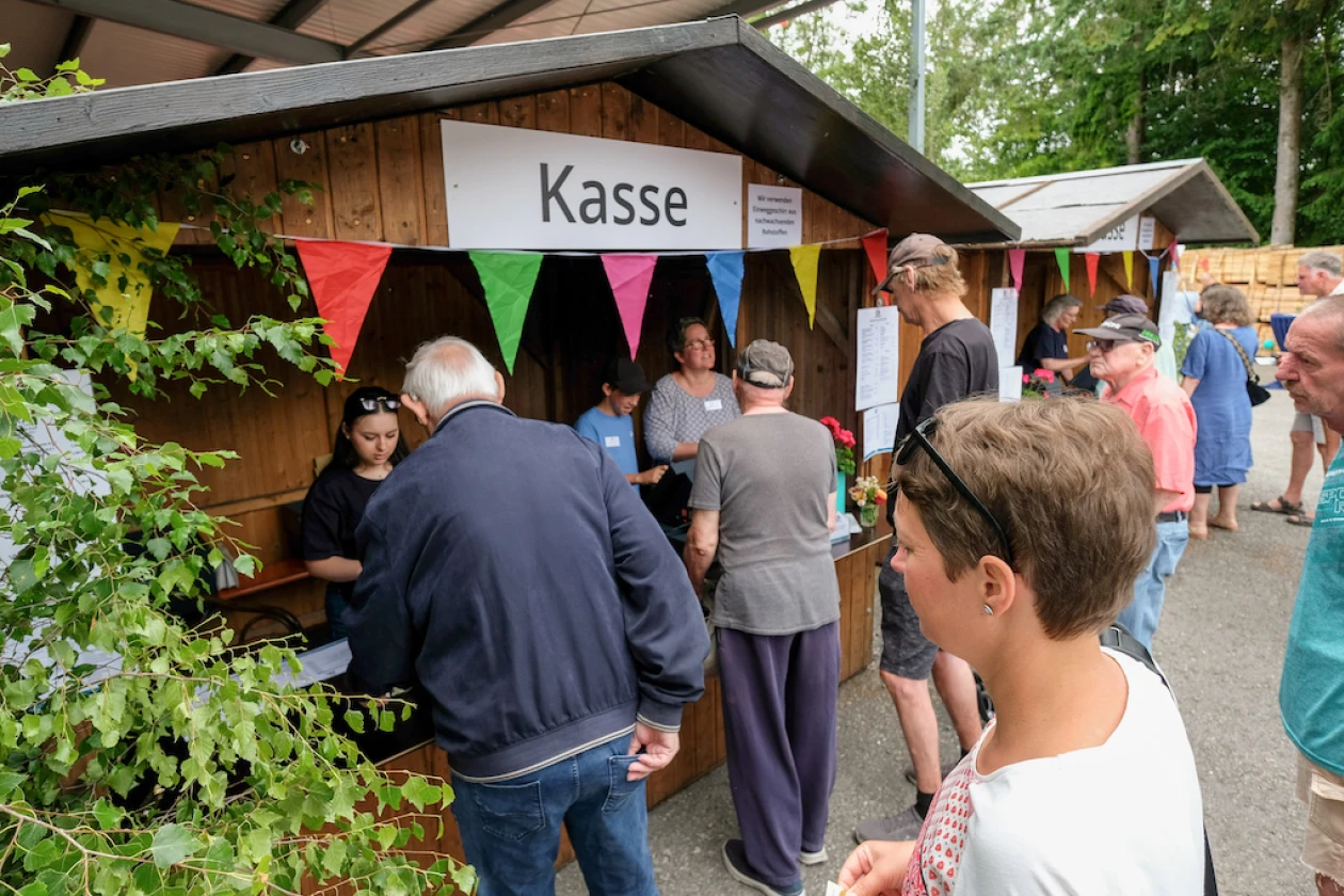 Mehrere Besucher stehen an einem Kassenh&auml;uschen mit bunten Wimpeln an. Das Holzgeb&auml;ude ist mit einem "Kasse"-Schild gekennzeichnet und von gr&uuml;nen Pflanzen umgeben.