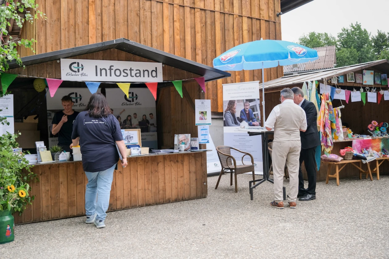 An einem Infostand mit Holzverkleidung und blauem Schirm informieren sich Besucher &uuml;ber die Arbeit der Erlacher H&ouml;he