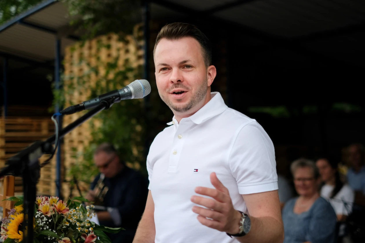 Ein Mann in wei&szlig;em Poloshirt spricht an einem Mikrofon vor Publikum. Er gestikuliert mit seiner Hand w&auml;hrend seiner Rede