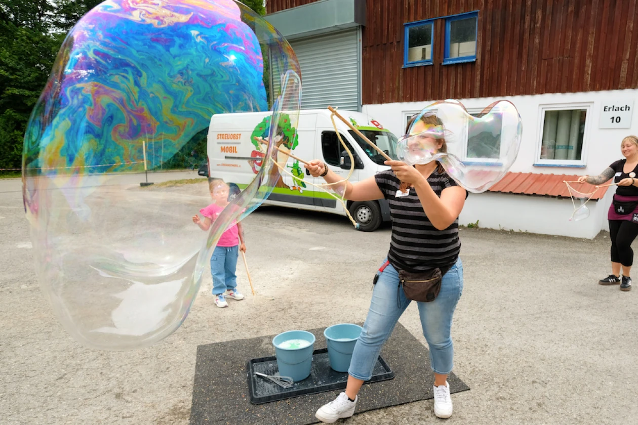 Eine Frau bl&auml;st mit einem Werkzeug eine riesige Seifenblase auf einem Platz auf, w&auml;hrend ein kleines Kind daneben steht und zuschaut