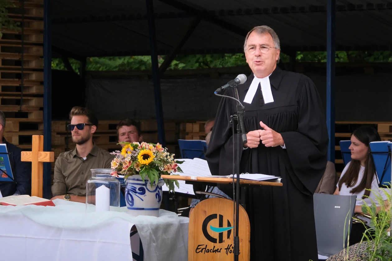 Ein Pastor in schwarzer Amtstracht spricht an einem Rednerpult w&auml;hrend einer Veranstaltung