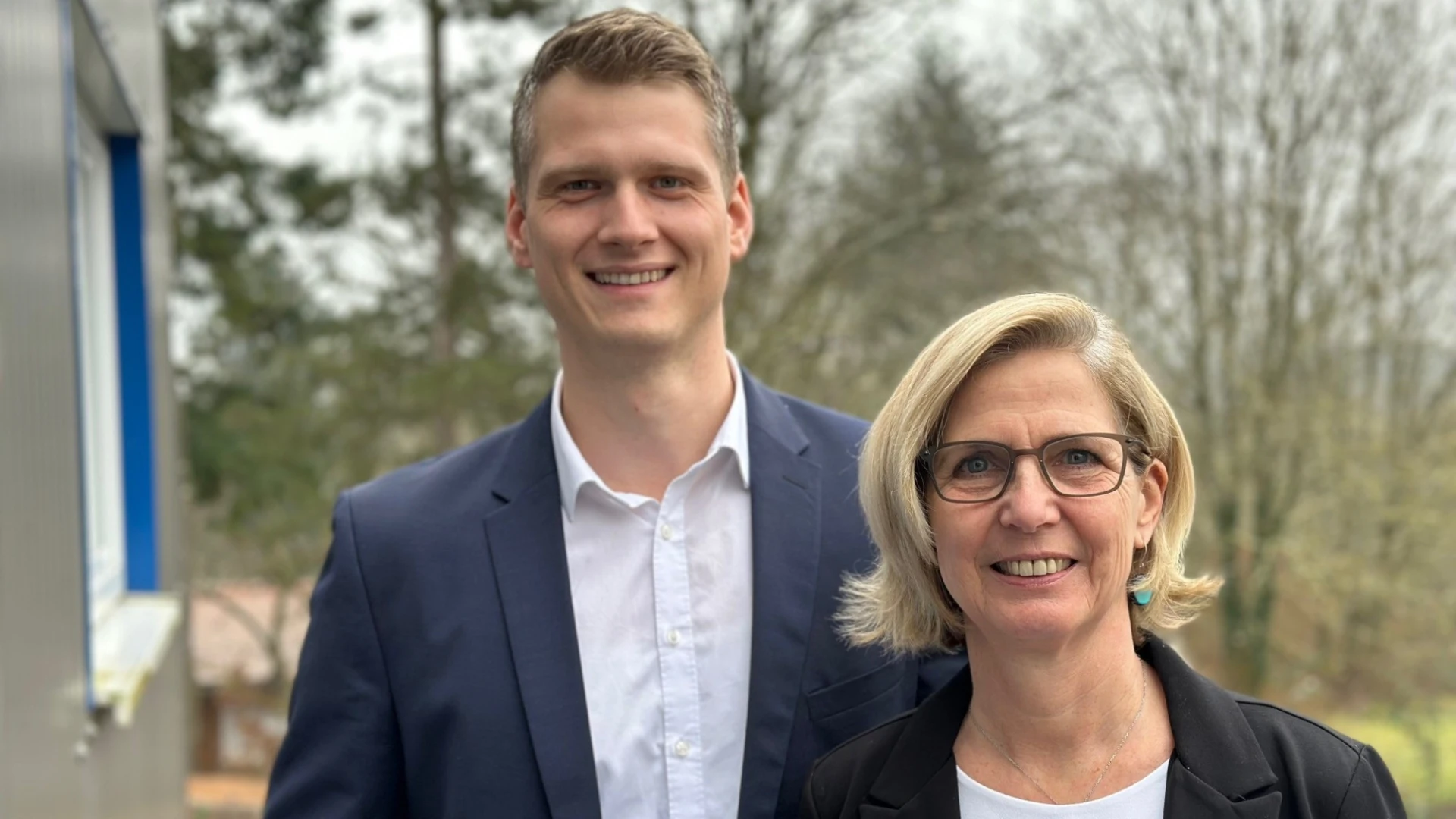 Ein junger Mann mit wei&szlig;em Hemd und blauem Sakko und eine blondhaarige Frau mit Brille, wei&szlig;em T-Shirt und schwarzen Blazer stehen neben einem Geb&auml;ude. Im Hintergrund sind B&auml;ume zu sehen.