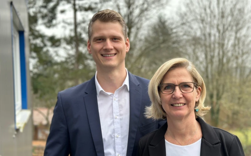 Ein junger Mann mit wei&szlig;em Hemd und blauem Sakko und eine blondhaarige Frau mit Brille, wei&szlig;em T-Shirt und schwarzen Blazer stehen neben einem Geb&auml;ude. Im Hintergrund sind B&auml;ume zu sehen.