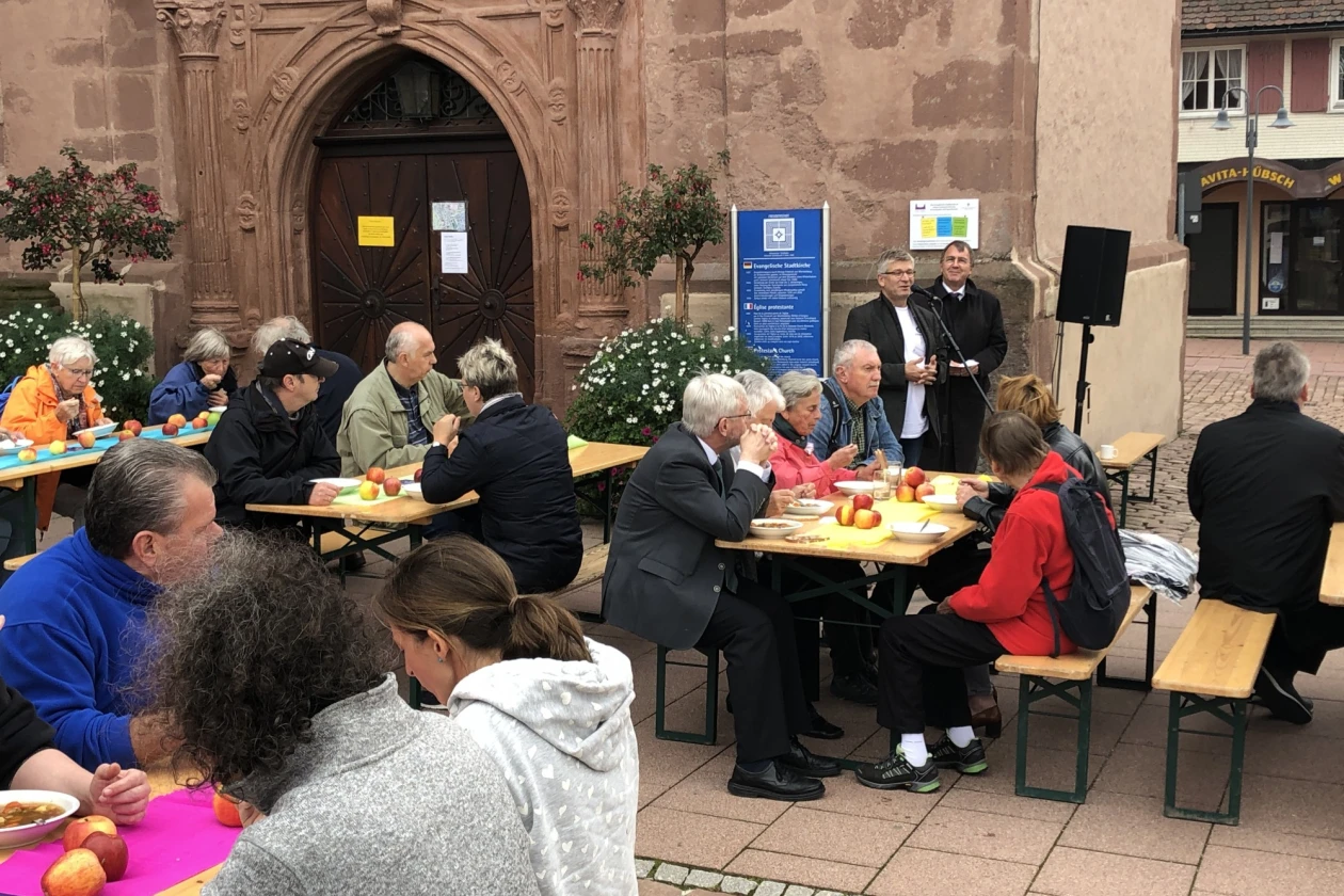 Gruppen von Menschen, die drau&szlig;en an Biertischgarnituren sitzen. Die Tische sind eingedeckt und mit &Auml;pfeln dekoriert. Die meisten Menschen haben einen Teller vor sich stehen. Im Hintergrund ein historisches Geb&auml;ude mit einer gro&szlig;en Holzt&uuml;r zu sehen. Rechts stehen zwei M&auml;nner hinter einem Mikrofon und einem Lautsprecher.
