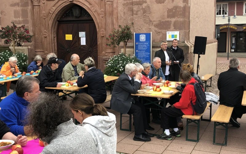 Gruppen von Menschen, die drau&szlig;en an Biertischgarnituren sitzen. Die Tische sind eingedeckt und mit &Auml;pfeln dekoriert. Die meisten Menschen haben einen Teller vor sich stehen. Im Hintergrund ein historisches Geb&auml;ude mit einer gro&szlig;en Holzt&uuml;r zu sehen. Rechts stehen zwei M&auml;nner hinter einem Mikrofon und einem Lautsprecher.