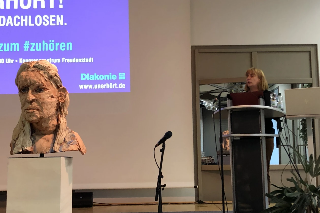 Eine Frau steht auf der B&uuml;hne hinter einem Rednerpult. Hinter ihr ist eine Leinwand zu sehen, die jedoch nur zur H&auml;lfte im Bild erscheint. Im Vordergrund befindet sich eine B&uuml;ste.