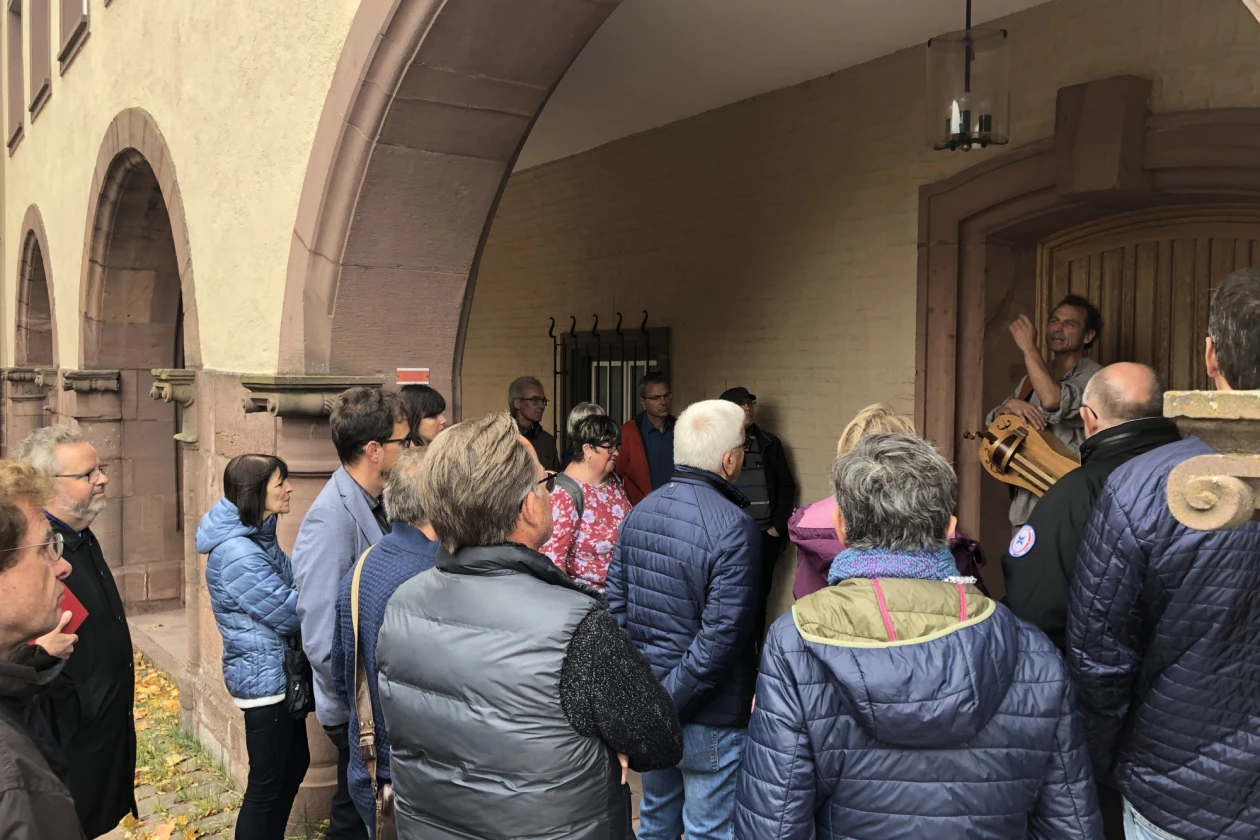 Auf dem Bild ist eine Gruppe von Menschen zu sehen, die in einem Torbogen vor einer Holzt&uuml;r stehen. Die Personen h&ouml;ren einem Mann zu, der etwas erkl&auml;rt und dabei ein Musikinstrument in der Hand h&auml;lt.