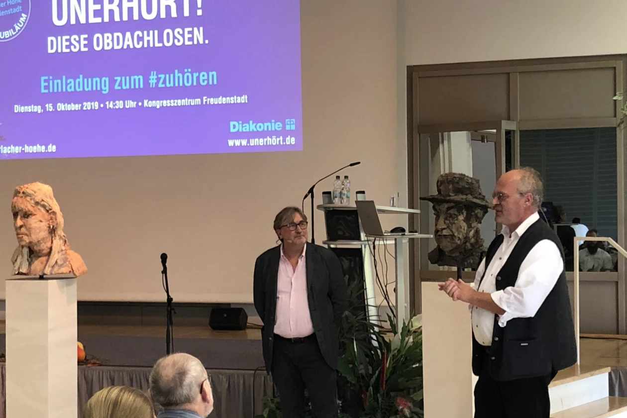 Zwei M&auml;nner stehen vor einer B&uuml;hne mit einer Leinwand, auf der die Aufschrift &sbquo;UNERH&Ouml;RT &ndash; Diese Obdachlosen. Einladung zum Zuh&ouml;ren.&lsquo; zu lesen ist. Links und rechts sind B&uuml;sten zu sehen. Im Vordergrund ragen die K&ouml;pfe zweier Zuschauer ins Bild.