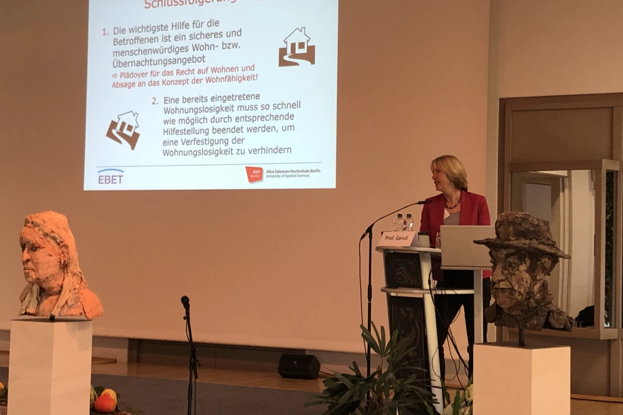 Eine Frau steht an einem Rednerpult auf der B&uuml;hne. Hinter ihr ist eine Leinwand mit dem Titel "Schlussfolgerungen" zu sehen. Links und rechts vor der B&uuml;hne stehen zwei B&uuml;sten.