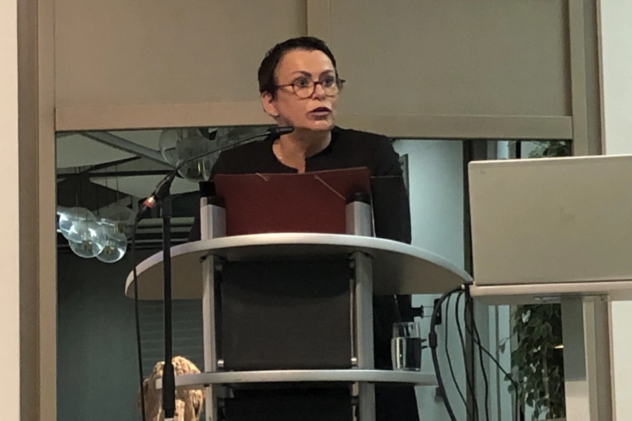 Eine dunkelhaarige Frau mit Brille steht hinter einem Rednerpult. Rechts neben ihr befindet sich ein Laptop auf einem Stehtisch. Die Frau tr&auml;gt schwarze Kleidung.