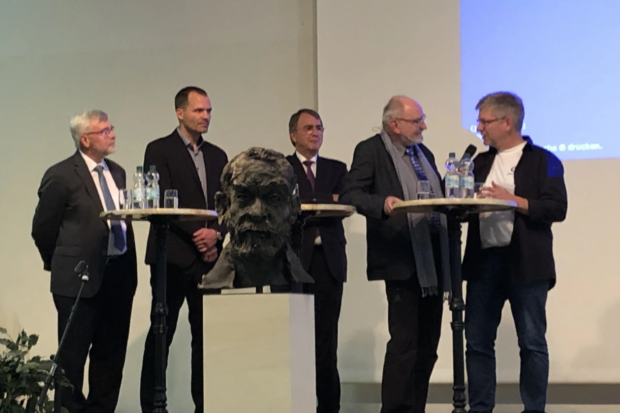 F&uuml;nf M&auml;nner stehen an einem Podium mit Stehtischen, auf denen Wasserflaschen und Gl&auml;ser stehen. Im Vordergrund ist die B&uuml;ste eines Mannes zu sehen. Die M&auml;nner tragen schwarze Anz&uuml;ge.