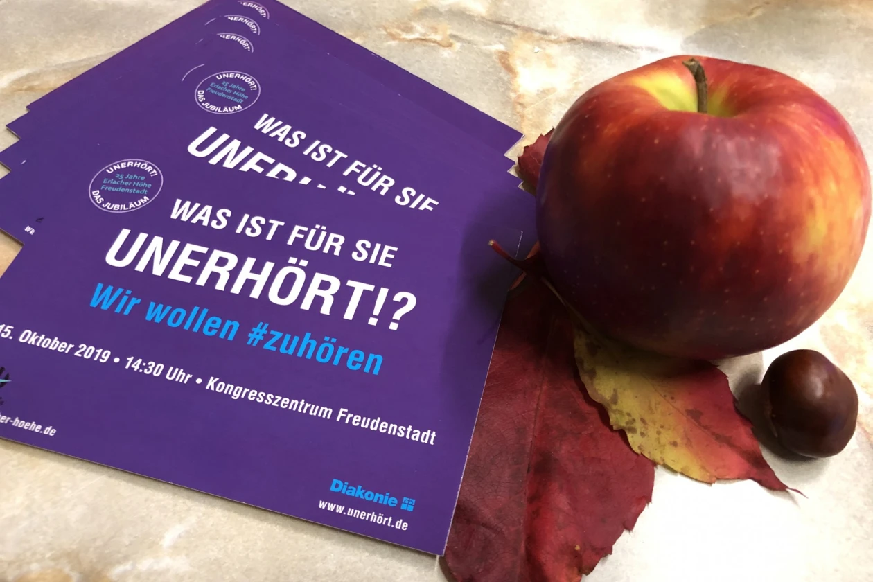 Lila Veranstaltungsflyer mit der Aufschrift: "Was ist f&uuml;r Sie unerh&ouml;rt?" neben einem roten Apfel auf herbstlichen Laub neben einer Kastanie.