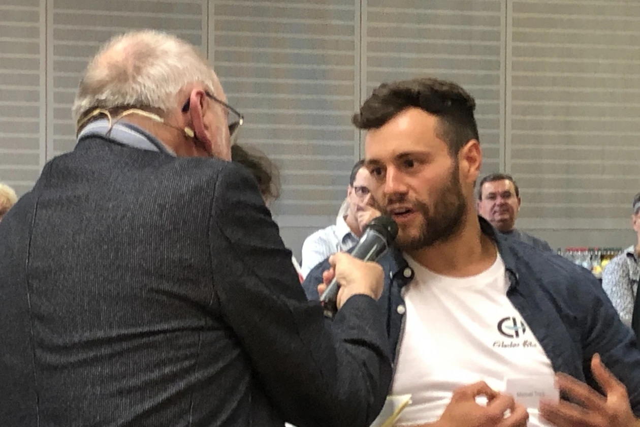 Der Mann links h&auml;lt einem Mann ein Mikrofon vor den Mund. Im Hintergrund sind weitere Personen zu sehen.