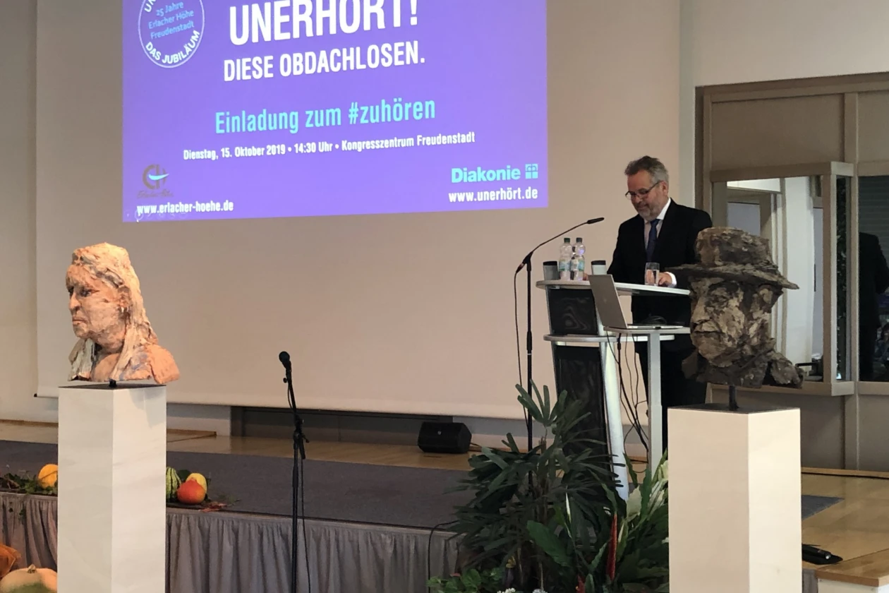 Ein Mann steht an einem Rednerpult auf einer B&uuml;hne. Hinter ihm ist eine Leinwand mit der Aufschrift "UNERH&Ouml;RT"! Diese Obdachlosen" zu sehen. Es ist eine Einladung zum Zuh&ouml;ren. Links und rechts vor der B&uuml;hne sind B&uuml;sten zu sehen.