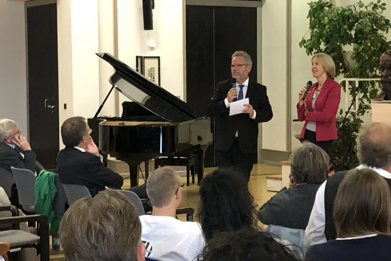 Ein &auml;lterer Mann und eine Frau stehen mit Mikrofon vor sitzenden Personen, die gespannt zuh&ouml;ren.