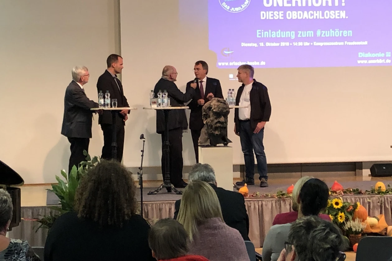 F&uuml;nf Personen stehen auf einer B&uuml;hne an Stehtischen vor einer Leinwand mit der Aufschrift Diese Obdachlosen - Einladung zum zuh&ouml;ren. Im Vordergrund sitzt Publikum und die B&uuml;ste eines Mannes ist vor der B&uuml;hne zu sehen.