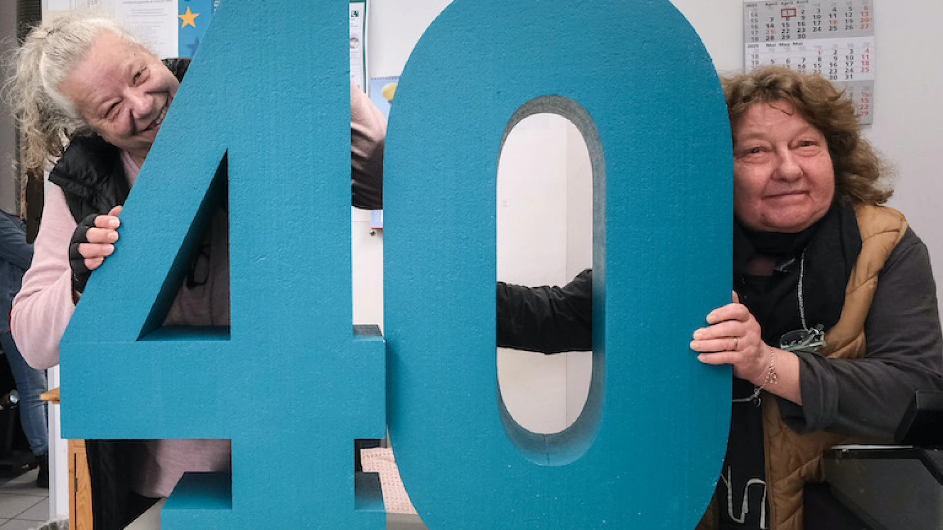 Zwei Frauen halten eine gro&szlig;e t&uuml;rkisfarbene Nummer "40" in einem B&uuml;ro, im Hintergrund ist ein Kalender f&uuml;r 2025 zu sehen.