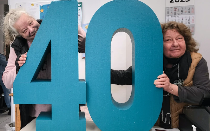 Zwei Frauen halten eine gro&szlig;e t&uuml;rkisfarbene Nummer "40" in einem B&uuml;ro, im Hintergrund ist ein Kalender f&uuml;r 2025 zu sehen.