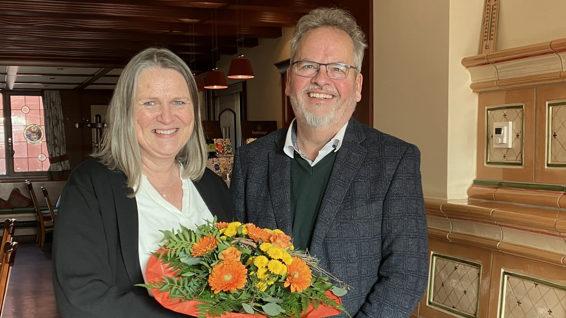 Zwei Personen stehen in einem Raum, beide l&auml;cheln in die Kamera. Die Frau mit wei&szlig;er Bluse und schwarzer Jacke h&auml;lt einen Blumenstrau&szlig; in der Hand, der Mann rechts tr&auml;gt ein graues Jackett, darunter einen dunklen Pullover und eine Brille.