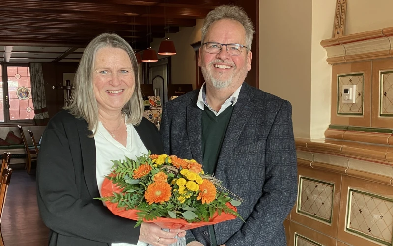 Zwei Personen stehen in einem Raum, beide l&auml;cheln in die Kamera. Die Frau mit wei&szlig;er Bluse und schwarzer Jacke h&auml;lt einen Blumenstrau&szlig; in der Hand, der Mann rechts tr&auml;gt ein graues Jackett, darunter einen dunklen Pullover und eine Brille.