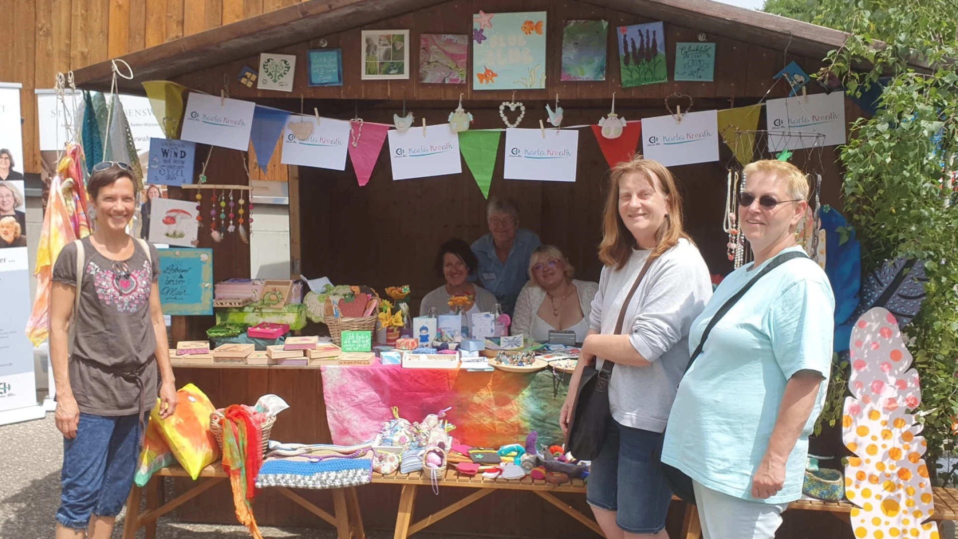 Vier Frauen pr&auml;sentieren an einem bunten Infostand mit handgemalten Kunstwerken und Dekorationen ihre kreativen Werke beim Jahresfest der Erlacher H&ouml;he