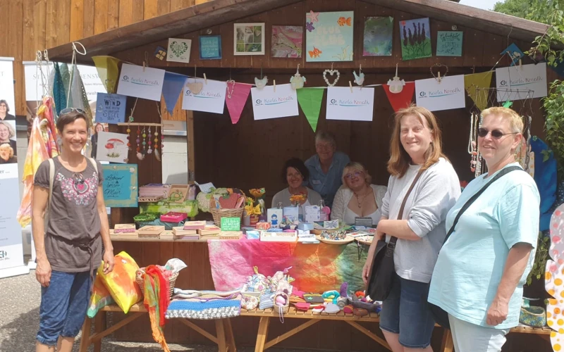 Vier Frauen pr&auml;sentieren an einem bunten Infostand mit handgemalten Kunstwerken und Dekorationen ihre kreativen Werke beim Jahresfest der Erlacher H&ouml;he