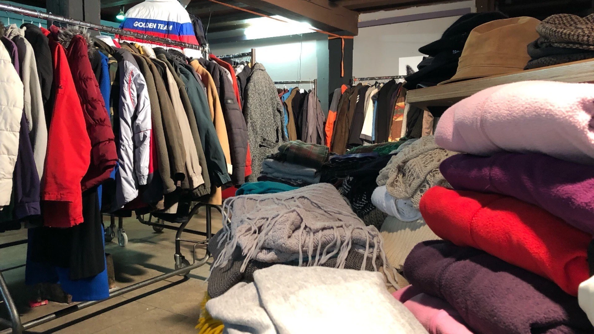 Ein Lagerraum mit gespendeten Winterkleidungsst&uuml;cken, darunter M&auml;ntel, Jacken und Pullover auf Kleiderstangen und Stapeln