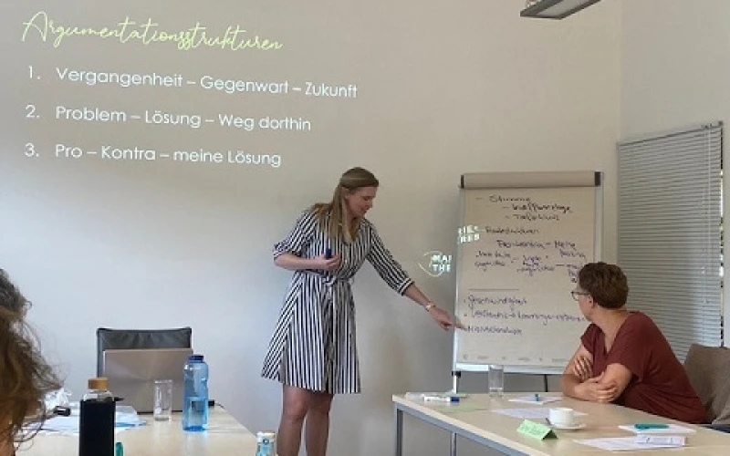 Eine Frau mit blonden Haaren und schwarz-wei&szlig; gestreiften Kleid steht vor einer Gruppe Menschen an einem Flipchart. Mit einem Beamer sind Schulungsinhalte an eine Wand projiziert.
