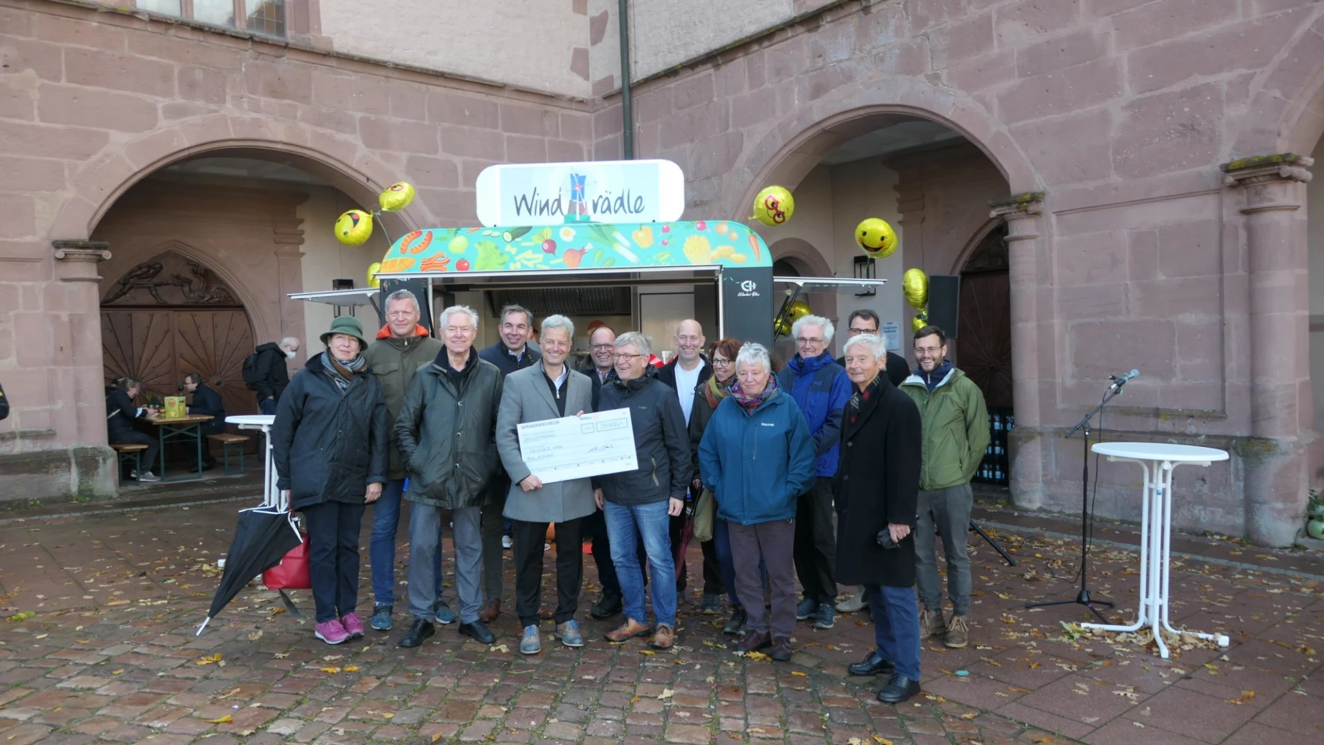 Mehrere Personen stehen vor einem Imbisswagen mit dem Schild "Windr&auml;dle". Der Mann in der Mitte h&auml;lt einen Scheck in der Hand.