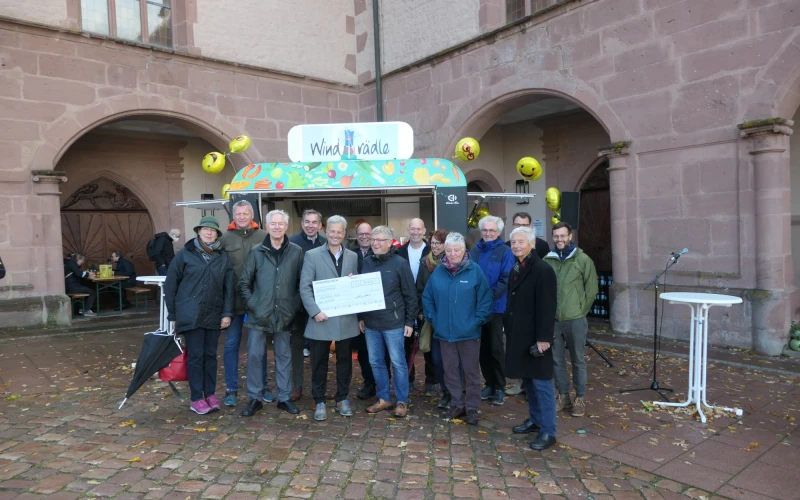 Mehrere Personen stehen vor einem Imbisswagen mit dem Schild "Windr&auml;dle". Der Mann in der Mitte h&auml;lt einen Scheck in der Hand.