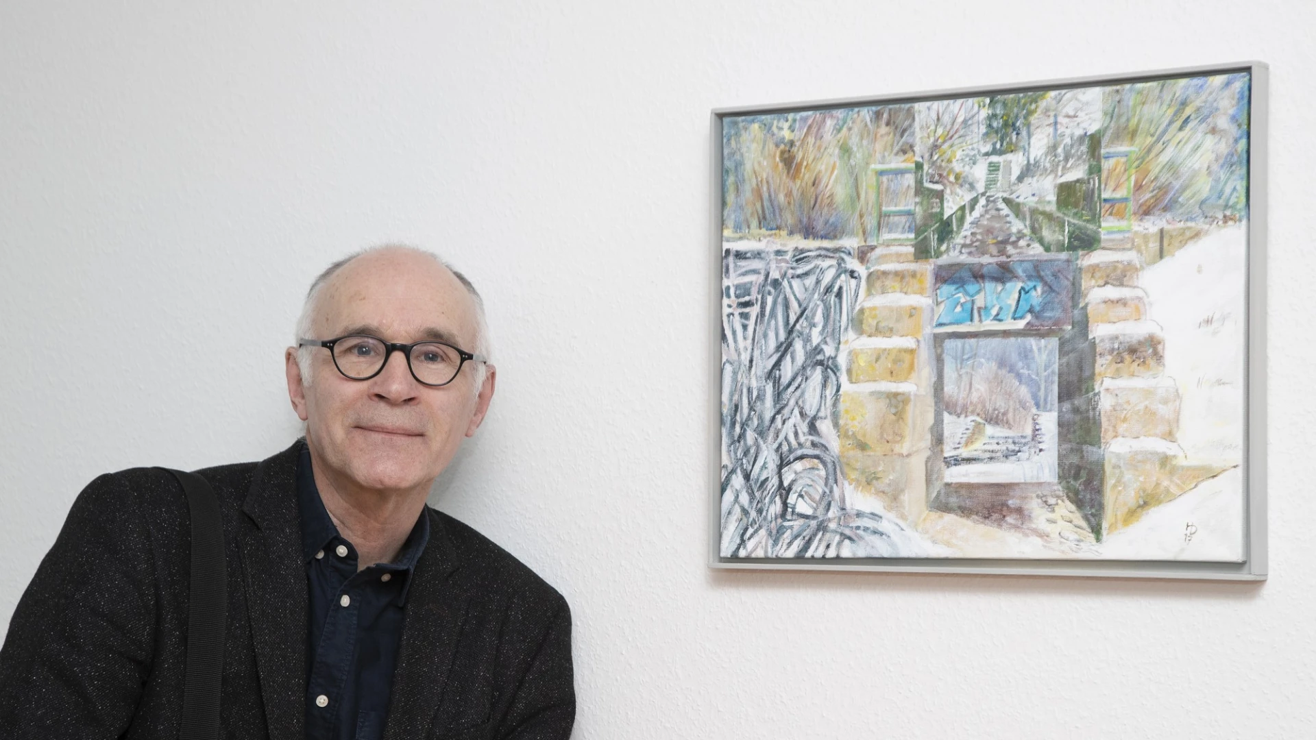 Ein &auml;lterer schwarz gekleideter Mann mit Brille steht neben einem abstrakten Kunstgem&auml;lde an einer wei&szlig;en Wand. Er l&auml;chelt und blickt direkt in die Kamera.