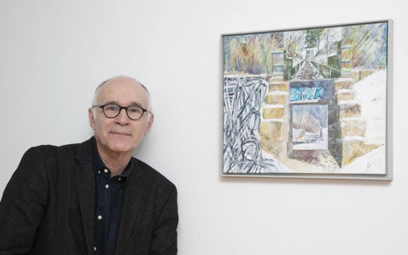 Ein &auml;lterer schwarz gekleideter Mann mit Brille steht neben einem abstrakten Kunstgem&auml;lde an einer wei&szlig;en Wand. Er l&auml;chelt und blickt direkt in die Kamera.