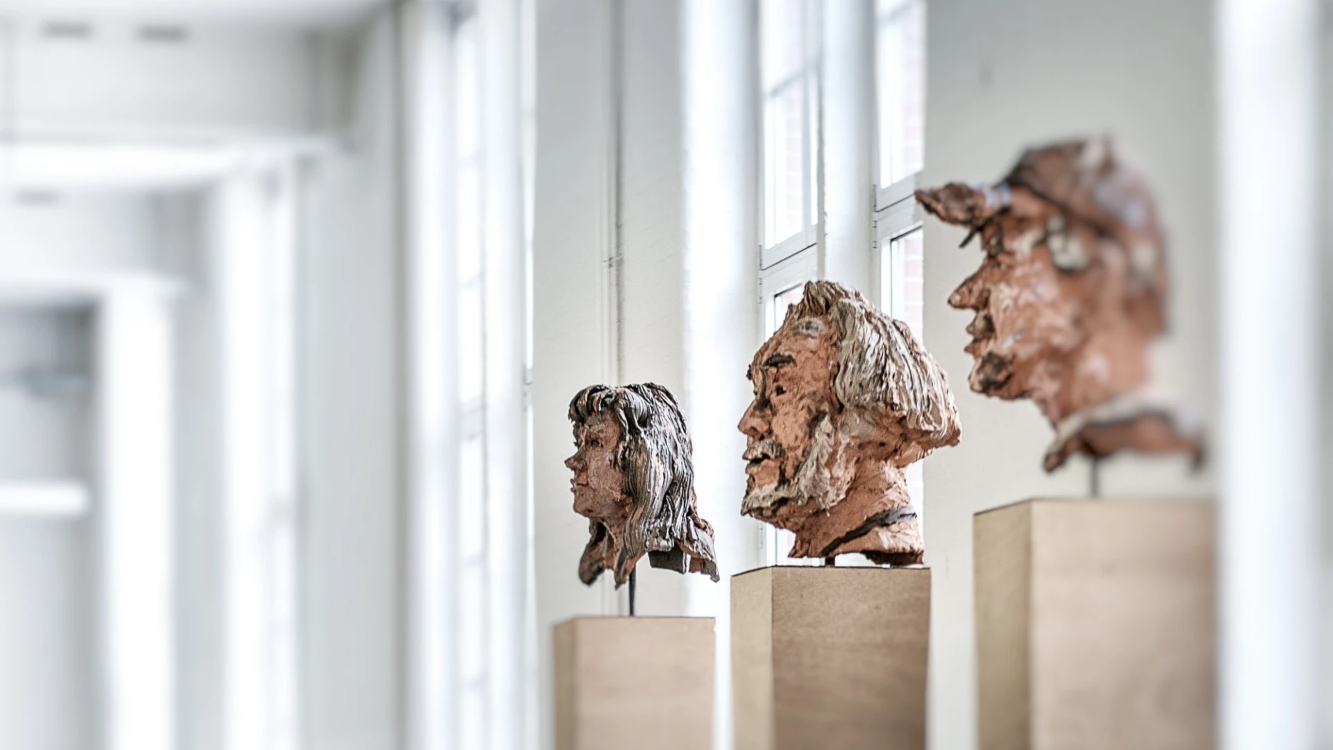 Drei Kopfskulpturen aus Ton sind zu sehen, auf h&ouml;lzernen Sockeln, die in einem hellen Raum mit Fenstern ausgestellt sind. Die Skulpturen zeigen verschiedene Gesichtsz&uuml;ge und Gesichtsausdr&uuml;cke.