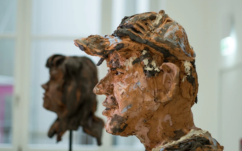Zwei Kopfskulpturen aus Ton, die ein menschliches Gesicht darstellt. Die Skulptur steht vor einem unscharfen Hintergrund mit einem Fenster.